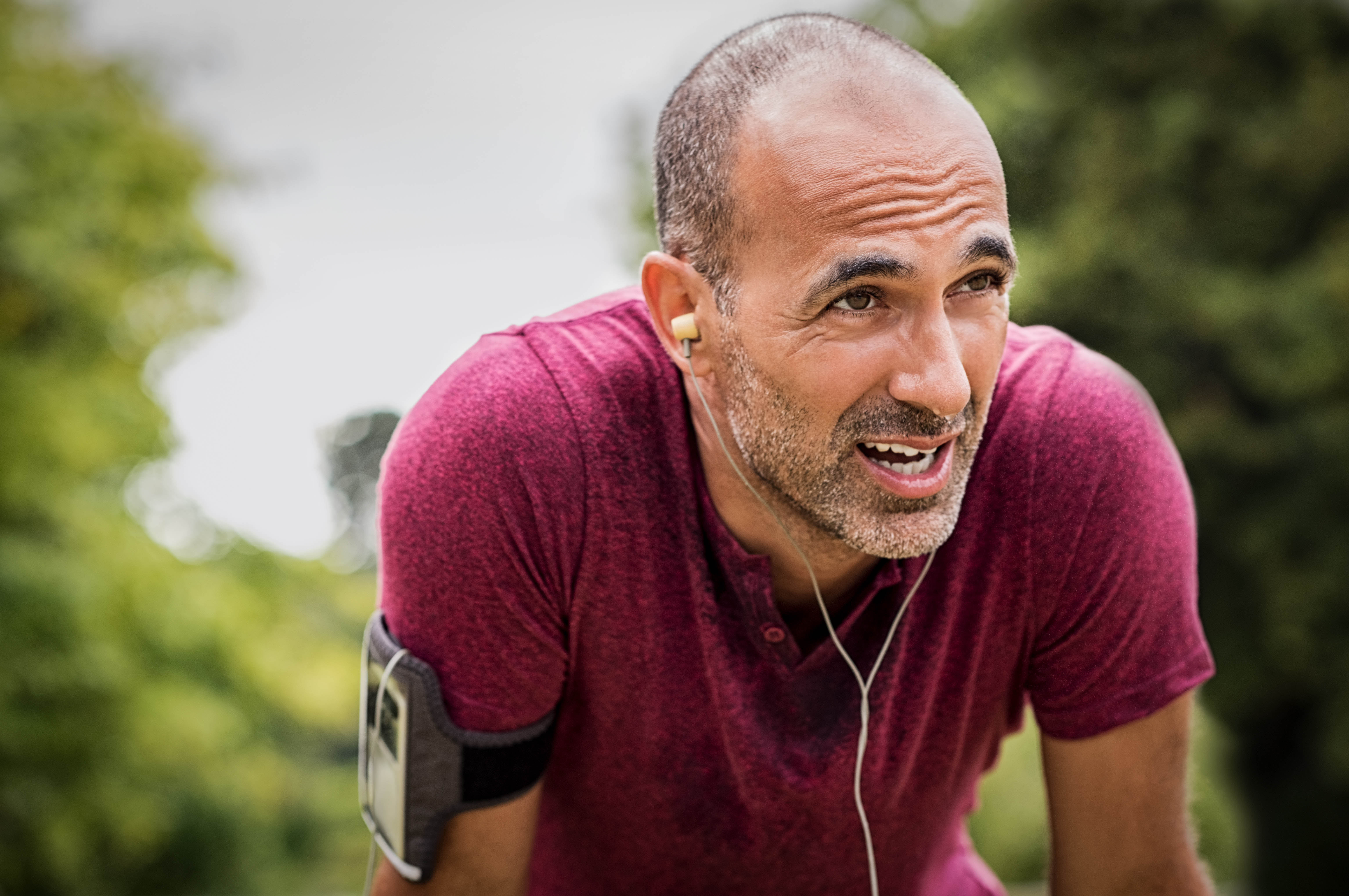 Middle aged man in a red t-shirt, hands on his knees, sweat dripping down his face, headphones in his ear, phone in a case on his arm